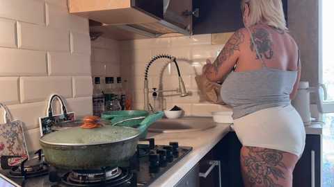 A video of a tattooed woman in a gray tank top and white shorts, cooking in a modern kitchen with stainless steel appliances, white subway tiles, and a large green pot on the stove.