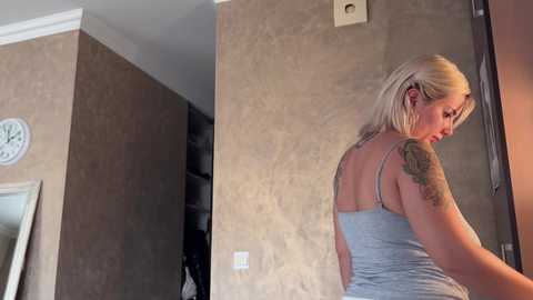 A video shows a blonde woman with a tattooed shoulder, wearing a light gray tank top, standing indoors near a doorway. The background features a beige textured wall with a clock and a partially visible mirror.