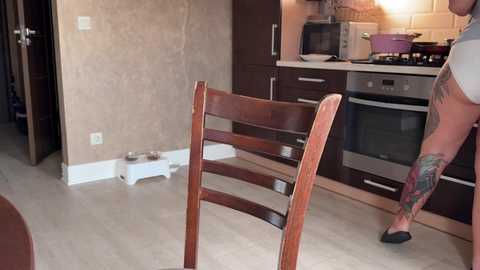 A video of a modern kitchen featuring a tattooed man in a white shirt and black slippers, standing next to a wooden chair. The kitchen has dark wood cabinets, stainless steel appliances, and beige tiled walls.