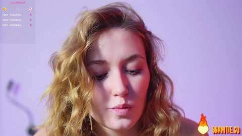 A close-up video of a fair-skinned, blonde woman with wavy hair, wearing subtle makeup, against a purple-lit background.
