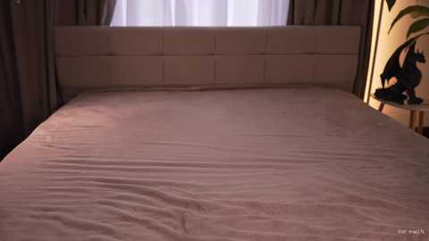 Video of a minimalist bedroom with a large, neatly made bed covered in a beige sheet, a white headboard, and a window with sheer curtains.