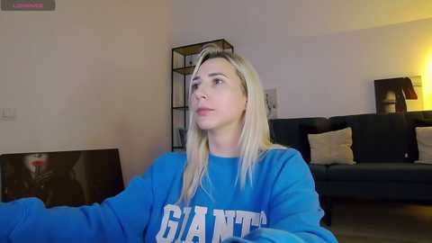 Video of a blonde woman in a blue \"Giant\" sweatshirt, sitting indoors. Background features a dark sofa, bookshelf, and a lamp.