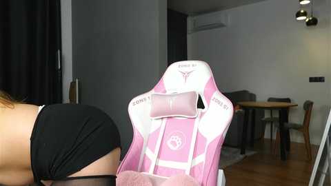 Video of a person in a black top leaning on a pink and white gaming chair with a pink pillow, in a modern, minimalist living room with a wooden floor, white walls, and an air conditioning unit.