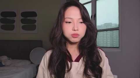 Video of a young East Asian woman with long, wavy black hair, wearing a beige sweater, sitting in a dimly lit bedroom with a window and modern art on the wall.