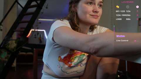 Video of a smiling woman with curly hair wearing a T-shirt, sitting at a desk in a dimly lit room with a staircase and gaming console visible.