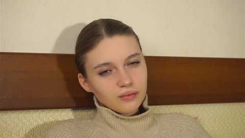Video of a young Caucasian woman with fair skin and straight, dark brown hair pulled back. She wears a cream-colored turtleneck sweater. The background is a beige wall with a wooden panel below.