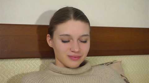 Video of a young woman with fair skin, brown hair tied back, wearing a beige turtleneck sweater, seated against a beige wall with a wooden panel above.