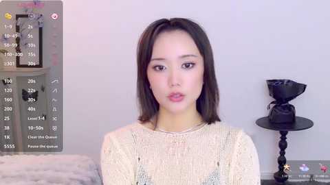 Video of a young East Asian woman with straight dark hair, fair skin, and light makeup. She wears a beige knit sweater. Background includes a white wall, a black candleholder, and a bed with a fluffy white blanket.