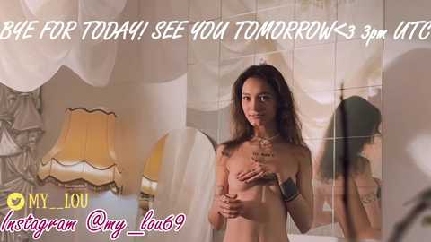 A video of a nude woman with medium breasts and light skin, holding a glass, standing in front of a mirror, in a room with a yellow lamp and white curtains.