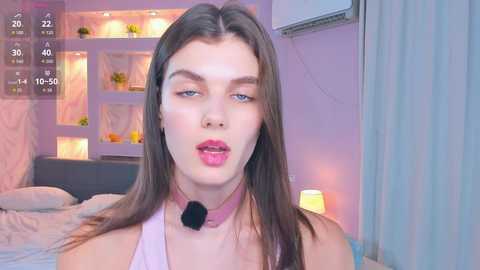 Video of a young woman with fair skin and long brown hair, wearing a pink choker and makeup, in a pastel-colored bedroom with a bed, shelves, and a lamp.