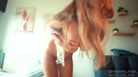 Video of a topless woman with long blonde hair, bending forward, wearing a white lace bra, in a modern, brightly-lit bedroom with minimalistic decor.