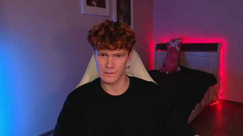 A video of a young man with curly red hair wearing a black t-shirt and a white, inflatable dragon neck brace, sitting on a bed with a red LED light strip, a cat, and a framed picture on a beige wall.