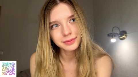 Video of a smiling, light-skinned young woman with long, straight blonde hair, blue eyes, and fair skin, in a softly lit, minimalist room with a metallic wall-mounted lamp.