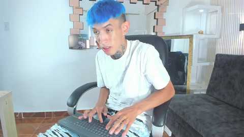 Video of a young, blue-haired person with a tattooed neck, wearing a white t-shirt and light blue jeans, playing a keyboard in a modern living room with white walls, a black chair, and a cat tree.