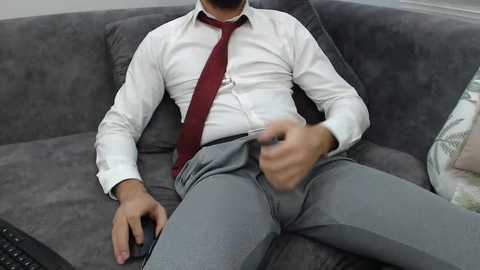 Video of a man in a white dress shirt, red tie, and gray pants, sitting on a gray couch, holding a remote, looking relaxed.