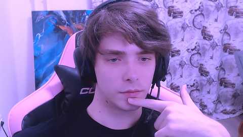 Video of a young Caucasian male with light brown hair, wearing headphones, looking contemplative, sitting in a pink gaming chair with a blue and brown abstract painting behind him.