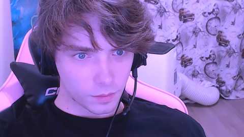 Video of a young man with tousled brown hair, wearing a black shirt and black headphones, sitting in a pink chair, with a cat toy and a white blanket in the background.