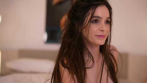 Media: Video of a smiling woman with long, wavy brown hair, wearing a strapless top, in a softly lit, beige bedroom.