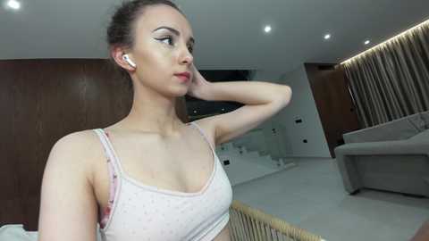 Video of a fair-skinned woman with brown hair in a ponytail, wearing a light pink spaghetti-strap top, standing in a modern, minimalist living room with white walls and wooden accents.