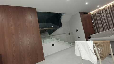 Video of a modern, minimalist living room with a wooden wall, glass staircase, white sofa, beige curtains, and a white towel draped over a chair.