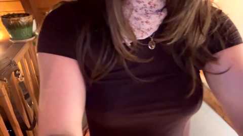 A video of a woman with long brown hair, wearing a black t-shirt and a white lace mask, standing in a rustic wooden room with a green candle and a wooden shelf in the background.