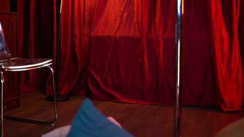 Video of a dimly lit room with a red curtain backdrop, a clear chair, and a blue garment on the floor.
