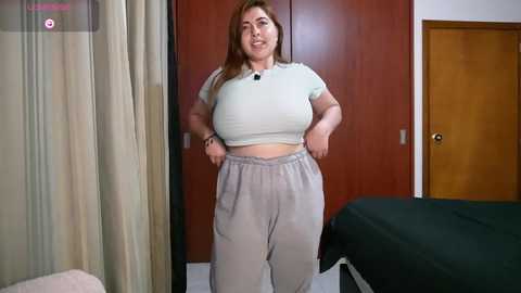Video of a plus-sized woman with light skin and long brown hair, wearing a tight white crop top and loose grey sweatpants, standing in a bedroom with wooden doors, a bed, and beige curtains.