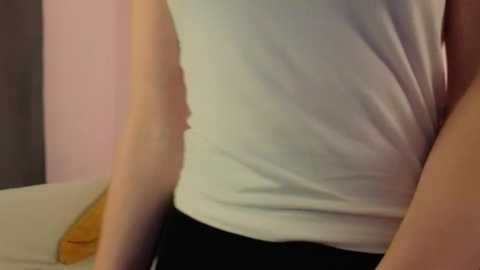 Video of a person's upper body, wearing a white tank top and black pants, sitting on a beige couch with a brown pillow on the left. The background shows a pink wall.