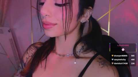 Video of a young woman with pale skin and dark hair styled in two braids, wearing a black top, nose ring, and several tattoos. She has a serene expression.