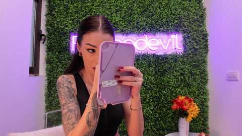 A tattooed woman with long dark hair, wearing a black top, takes a selfie in front of a vibrant green wall with \"fadedevil\" neon sign. She holds a pink smartphone with a white case.