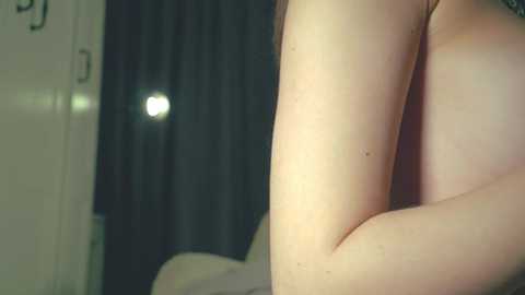 A video captures the side profile of a light-skinned person's arm, showing smooth skin and a small mole. The background features a dark curtain and a dimly lit room, suggesting an intimate, indoor setting.