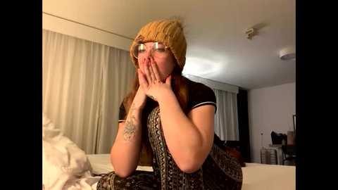 Video of a light-skinned woman with long brown hair, wearing a beige knit beanie, black t-shirt, and glasses, covering her face with her hands, in a dimly lit room with white curtains, cluttered furniture, and a patterned blanket.