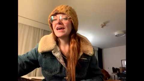 A video of a smiling, red-haired woman with glasses, wearing a beige knitted hat and a denim jacket with a fur collar, in a cozy, dimly lit room.