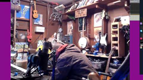 A video of a cluttered, cozy music room with a dark-haired person in a gray hoodie leaning on a keyboard, surrounded by guitars, drums, posters, and various musical instruments.
