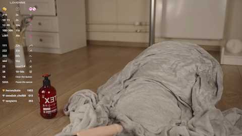 Video of a person wrapped in a gray blanket, lying on a light wooden floor in a room with white cabinets and a red bottle of lotion.