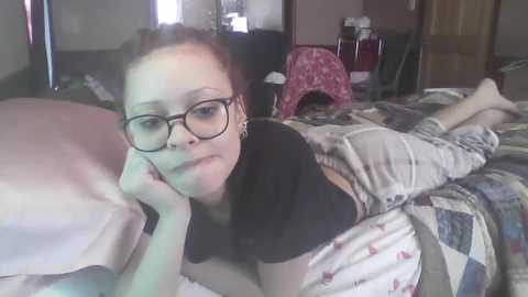 A video shows a young woman with light skin, glasses, and brown hair, lying on a quilted bed in a dimly lit bedroom, wearing a black crop top and beige shorts.