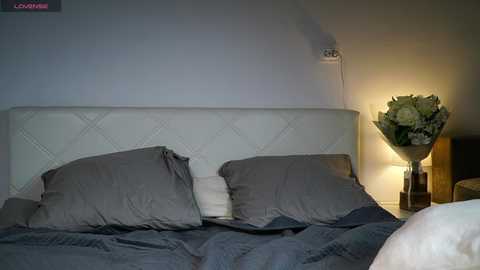 Video of a minimalist bedroom with a white, tufted headboard, grey pillows, and a grey duvet, featuring a bedside table with a vase of white flowers and a lit lamp, against a plain white wall.