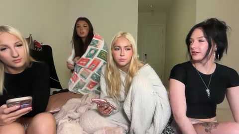Video of four young women, two Asian and two Caucasian, lounging on a beige couch, one with a blanket, another wearing a black crop top, all with playful expressions.
