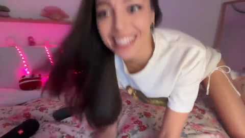 Video of a smiling, light-skinned woman with long black hair, wearing a white T-shirt and floral pajama shorts, lying on a bed with a floral sheet. Pink LED lights in the background.