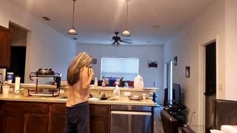 A video of a woman, topless with long blonde hair, in a modern kitchen with wooden cabinets, beige countertops, and stainless steel appliances. She's holding a camera, facing the window with blinds.