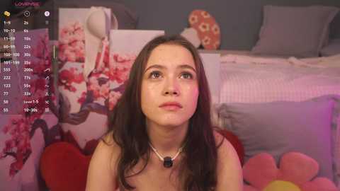 A video of a young woman with long brown hair, light skin, and a contemplative expression, sitting in a cozy bedroom with pink and floral decor, including a cherry blossom print and plush toys.