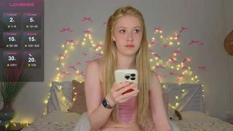 Media: A video of a young woman with long blonde hair taking a selfie in a cozy bedroom, adorned with fairy lights and pink bows on the wall, holding a white phone.