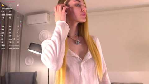 Video of a young woman with long blonde hair, wearing a white button-down shirt, adjusting her hair in a modern, minimalist room with a lamp, AC unit, and grey curtain.