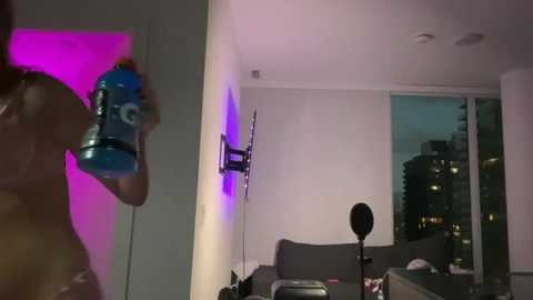 Video of a woman in a modern apartment, holding a blue water bottle, illuminated by purple light, with a cityscape visible through large windows.