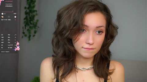Video of a young, fair-skinned woman with wavy, shoulder-length brown hair, wearing a silver necklace, in a neutral-toned room with a green plant.