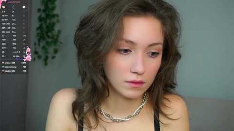 Video of a young woman with wavy brown hair, wearing a black top and silver necklace, looking introspective, in a soft-lit room with a green wall and a plant.