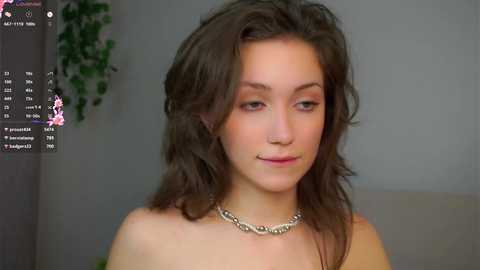 Video of a young Caucasian woman with shoulder-length wavy brown hair, wearing a silver chain necklace, and a subtle makeup look. She has a calm expression and is indoors, possibly on a couch, with a blurred background.