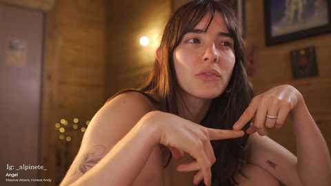 Video of a nude, fair-skinned woman with long dark hair, expressionless face, in a dimly lit, rustic room with wooden walls.