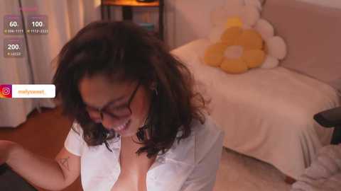 Media: Video of a woman with curly dark hair, wearing glasses and a white blouse, sitting on a beige couch with a stuffed toy, in a cozy room with wooden flooring.