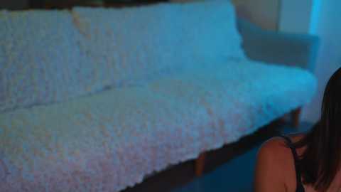 Video of a woman with long brown hair, wearing a black top, leaning forward on a textured, light blue sofa. The background is dimly lit, with a soft blue hue.